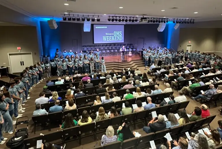 103 college students stand along the front of a worship center, facing people in pews being commissioned as summer missionaries.